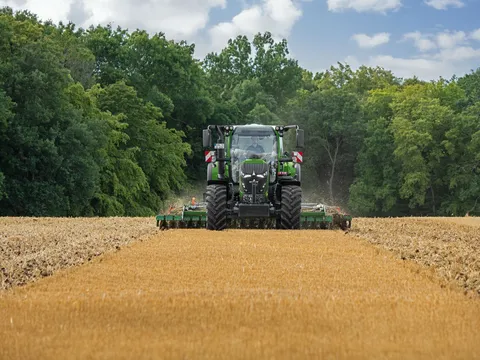 Ein grüner Fendt 620 Vario fährt mit einer Scheibenegge über ein Stoppelfeld im Hintergrund ein Wald mit grünen Bäumen.