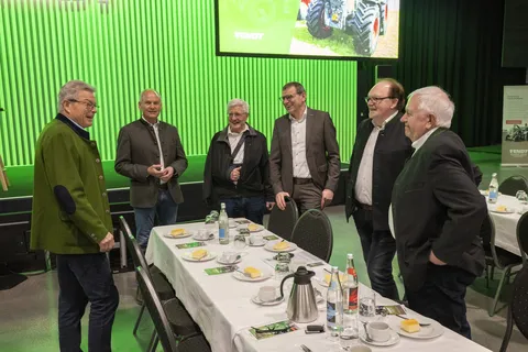 6 Männer stehen an einem gedeckten Tisch und unterhalten sich. Hinter ihnen befindet sich die Bühne mit grünem Hintergrund.