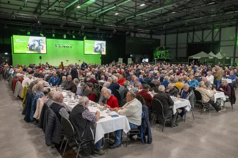 Menschen sitzen an gedeckten Tafeln und unterhalt sich. Neben ihnen befindet sich eine Bühne mit grünem Hintergrund und Fendt Logo sowie 2 befestigte Bildschirme.