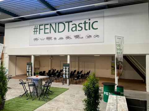 In einer Halle stehen 2 Stehtische mit Stühlen, dahinter Stuhlreihen unter einer Überdachung, die nach links zur Leinwand zeigen. Daneben stehen Broschürenständer und darüber hängt ein Schild mit grünem Schriftzug #FENDTastic und Produktsymbolen.