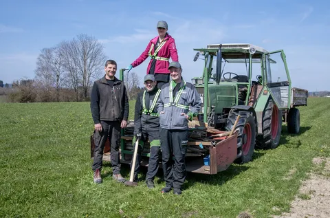 4 Personen, davon eine Frau, die auf den aufgeladenen Holzpfählen auf dem Fendt Traktor steht. Die 3 Männer stehen vor dem grünen Fendt Traktor.