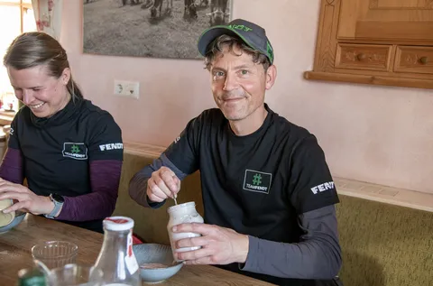 Eine Frau (links) und ein Mann (rechts) tragen Fendt Shirts und machen Mittagspause. Die Frau schaut lächelnd auf ihren Teller, während der Mann, der ein Glas Joghurt in der Hand hält, lächeln in die Kamera schaut.