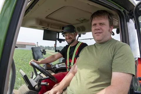 2 Männer sitzen in einem Traktor, dessen Innenraum zu sehen ist, und schauen in die Kamera. Einer sitzt am Lenkrad.