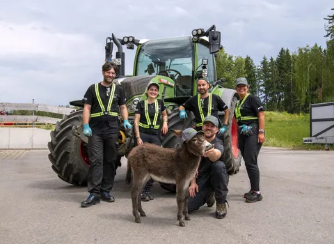 5 Personen, davon 2 Frauen stehen vor einem grünen Fendt Traktor und tragen Arbeitsschutzkleidung. Vor ihnen hockt ein Mann und füttert einen Esel mit Fendt Cap