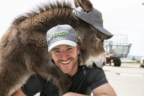 Ein Mann und ein Esel. Der Mann trägt ein schwarzes Shirt und ein Cap mit grünem Fendt Schriftzug. Er lächelt in die Kamera, während der Esel, der auch ein Fendt Cap trägt, ihm spielerisch in die Schulter beißt.