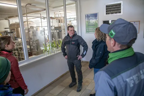 Menschen in einem Raum. In der Mitte des Fotos steht ein Mann, der eine Frau ansieht und ihr zuhört. Die Frau ist nur seitlich zu sehen. Um die beiden herum stehen weitere Menschen und schauen sie an.