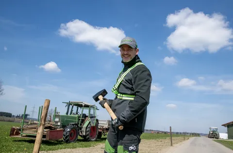 Ein Mann in Arbeitsschutzkleidung steht lächelnd vor einem Holzpfahl und hält einen Vorschlaghammer in der Hand. Links von ihm im Hintergrund steht ein grüner Fendt Traktor, auf dem weitere Pfähle liegen.