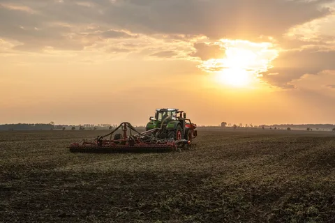 Der grüne Fendt 1000 Vario fährt mit einem Kverneland Fronttank und einer Scheibenegge über den Acker in Richtung Sonnenuntergang.