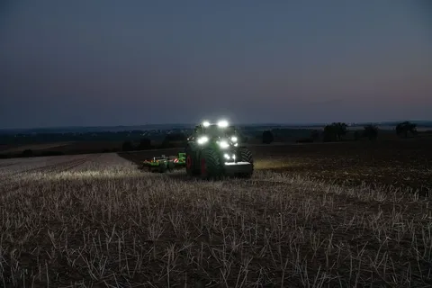 Ein grüner Fendt Traktor 1000 Vario bearbeitet mit einem Amazone Grubber in grün und orange einen Acker mit Stoppeln. Alle Scheinwerfer sind eingeschaltet, weil es dunkel ist.