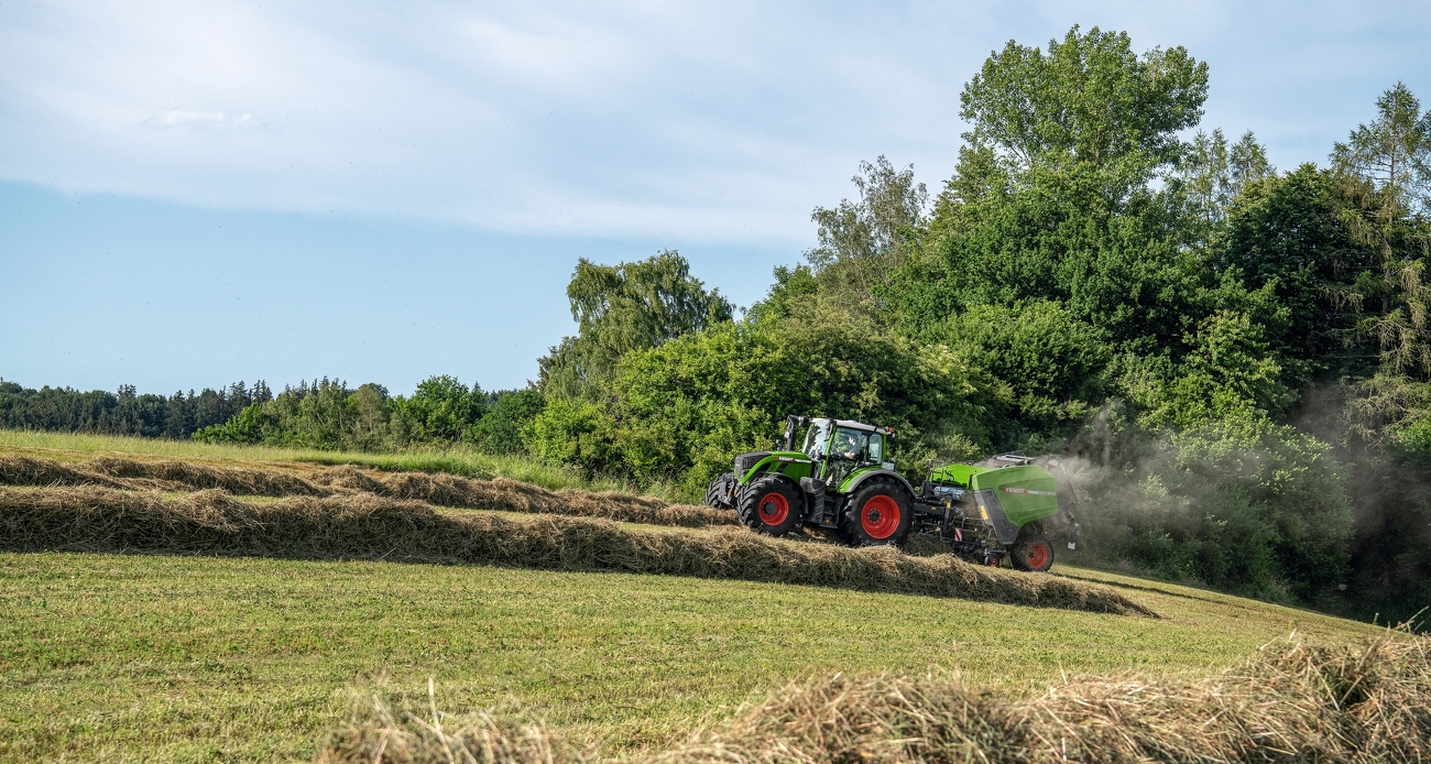 Fendt 700 Vario mit Rotana 180 V Xtra bei der Grünfütterernte. Im Hintergrund sind Bäume und Sträucher zu sehen.