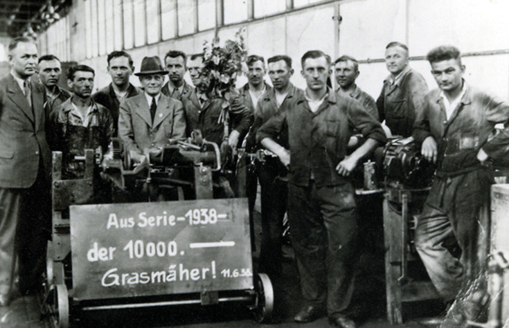 Schwarz weiß foto von Mitarbeitern von früher mit dem Schild: Aus Serie 1938 - der 10.000 Grasmäher