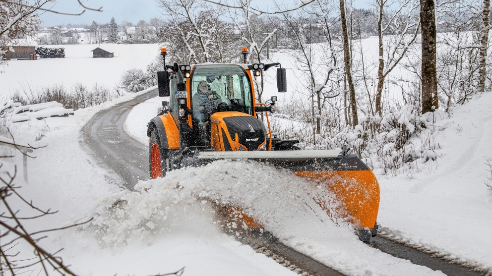 Orangener Fendt Traktor räumt Schnee auf einer Straße in einer verschneiten Landschaft