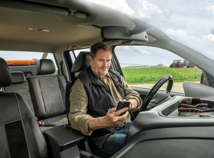 Landwirt sitzt im Auto auf sein Handy schauend mit Fendt Traktor im Hintergrund