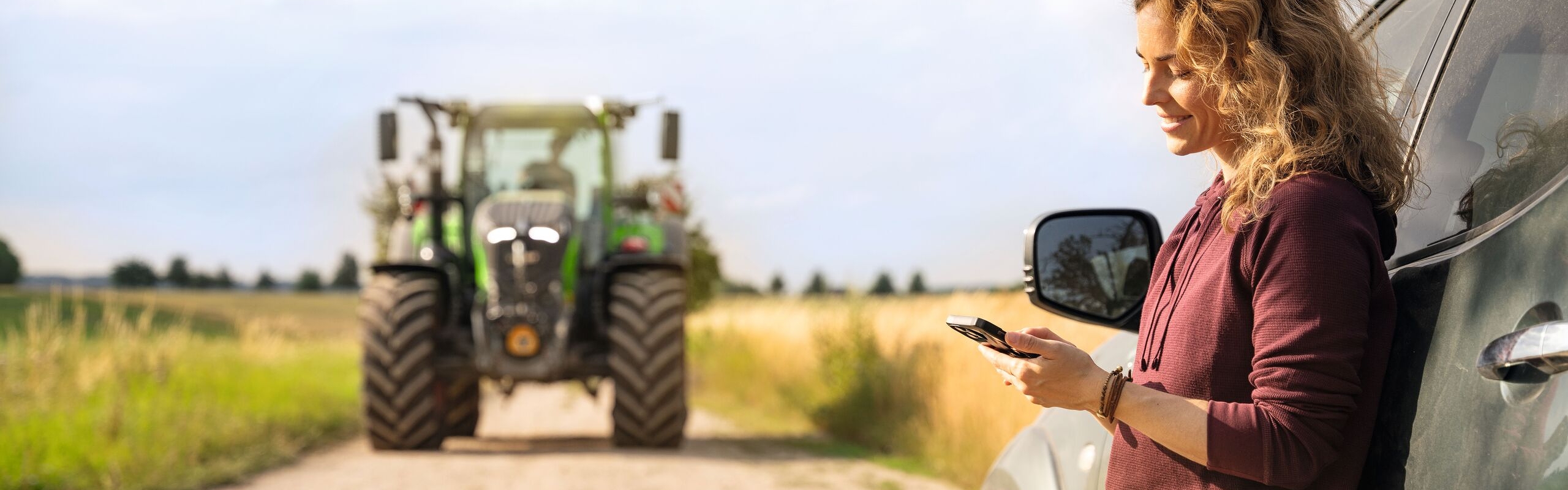 Eine Frau mit blonden Locken und dunkelroten Pulli steht angelehnt an einem Auto und schaut auf ihr Handy, im Hintergrund ist ein Fendt Traktor zu sehen