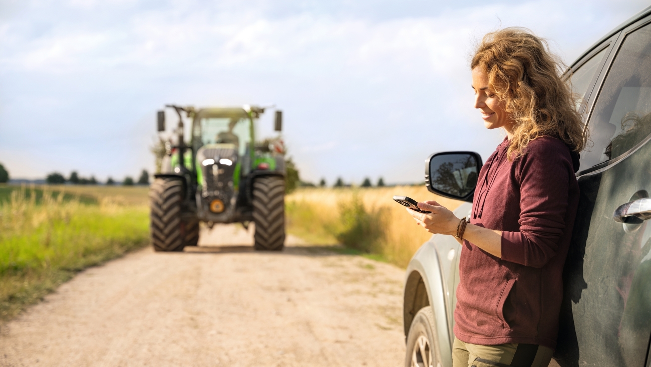 Eine Landwirtin lehnt sich an ihrem Auto an und liest den Fendt Newsletter auf dem Handy.