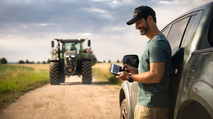 Ein Landwirt hält ein Tablet in der Hand und lehnt sich an sein Auto an. Im Hintergrund kommt ein Fendt 700 Vario Gen7 angefahren.