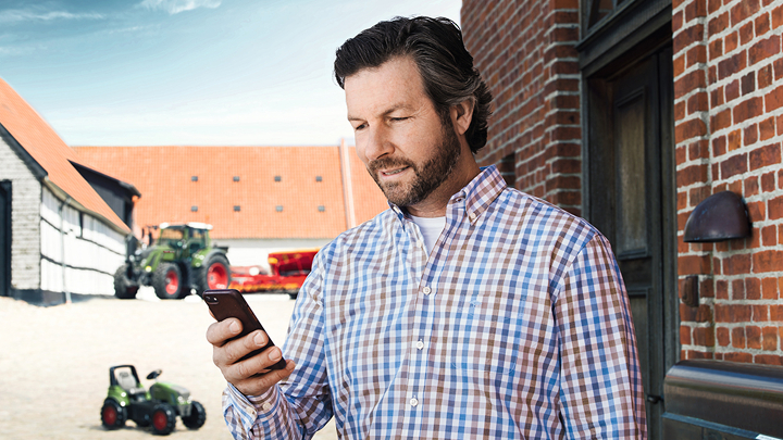 Ein Landwirt steht vor seinem Hof und hält ein Handy in der Hand.