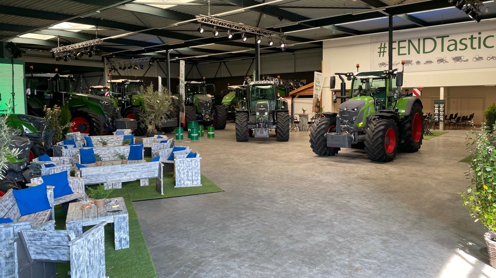 Händlerstandort Mechan in Achterveld, Niederlande, heute In einer Halle stehen Fendt Traktoren im Halbkreis. Links stehen Holztische und -stühle auf Rasenteppich. Hinter den Traktoren rechts stehen Stühle unter einer Überdachung und darüber hängt ein Schild mit grünem Schriftzug #FENDTastic und Produktsymbolen.
