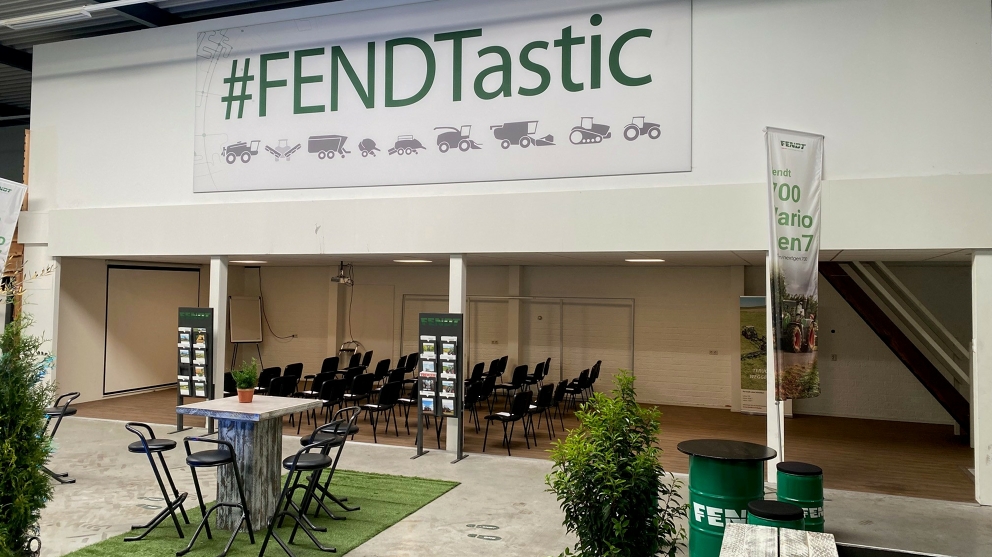 Händlerstandort Mechan in Achterveld, Niederlande, heute In einer Halle stehen 2 Stehtische mit Stühlen, dahinter Stuhlreihen unter einer Überdachung, die nach links zur Leinwand zeigen. Daneben stehen Broschürenständer und darüber hängt ein Schild mit grünem Schriftzug #FENDTastic und Produktsymbolen.