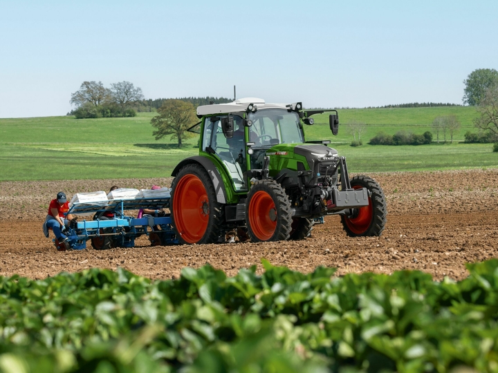 Der grüne Traktor Fendt e100 Vario mit roten Felgen fährt auf einem braunen Feld mit einem blauen Himmel und weißen Wolken im Hintergrund.