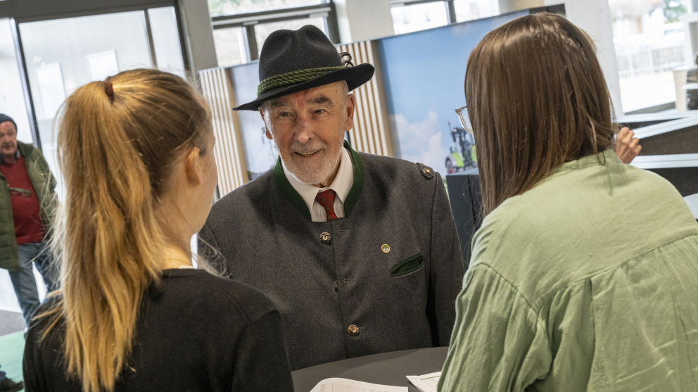 Begrüßung eines Gastes bei der Fendt Ehemaligenfeier 2025 im Fendt Forum Ein Mann mit Hut wird in der Eingangshalle von zwei Mitarbeiterinnen begrüßt, die mit dem Rücken zur Kamera stehen.