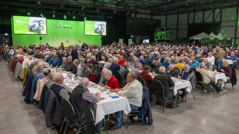 Ehemalige Fendt Mitarbeiter bei der Fendt Ehemaligenfeier 2025 im Fendt Forum Menschen sitzen an gedeckten Tafeln und unterhalt sich. Neben ihnen befindet sich eine Bühne mit grünem Hintergrund und Fendt Logo sowie 2 befestigte Bildschirme.