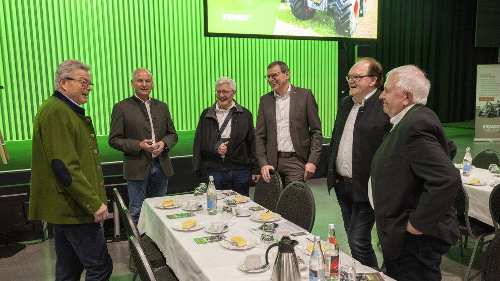 Sepp Nuscheler, Walter Wagner, Karl-Heinz Welz und Peter-Josef Paffen im Gespräch mit zwei weiteren Gästen der Fendt Ehemaligenfeier 2025. 6 Männer stehen an einem gedeckten Tisch und unterhalten sich. Hinter ihnen befindet sich die Bühne mit grünem Hintergrund.