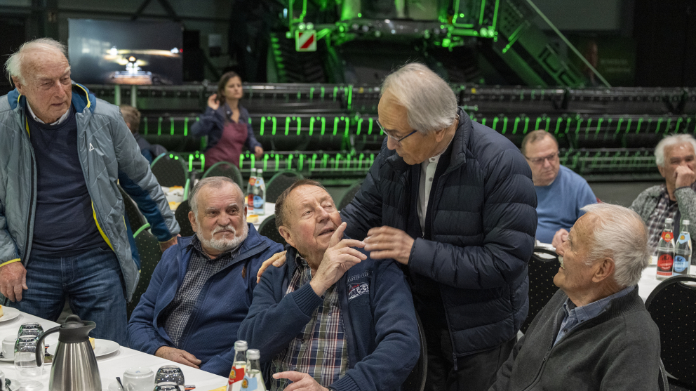 Ehemalige Fendt Mitarbeiter bei der Fendt Ehemaligenfeier 2025 im Fendt Forum 8 Menschen, im Hintergrund läuft eine Frau, 5 Männer sitzen und zwei stehen. Ein Mann hat seine Hand auf die Schulter eines anderen gelegt und unterhält sich mit ihm.
