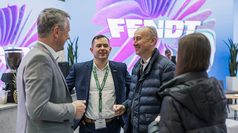 Georg Herbig spricht mit Fendt Kunden auf der Agrotech Kielce 2025 4 Menschen unterhalten sich, im Hintergrund ein großes weißes Fendt Logo auf buntem Hintergrund