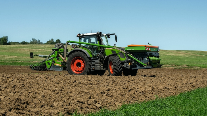 Der grüne Fendt 600 Vario mit roten Felgen und einer Drillmaschine als Anbaugerät fährt auf einem Acker. Im Hintergrund ist eine grüne Wiese und ein blauer Himmel zu sehen.