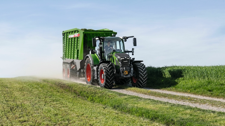 Der grüne Fendt 600 Vario Traktor mit roten Felgen und dem grünen Ladewagen Fendt Tigo fährt auf einer kleinen Feldstraße mit einer grünen Wiese im Vorder- und Hintergrund.