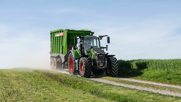 Der grüne Fendt 600 Vario Traktor mit roten Felgen und dem grünen Ladewagen Fendt Tigo fährt auf einer kleinen Feldstraße mit einer grünen Wiese im Vorder- und Hintergrund.