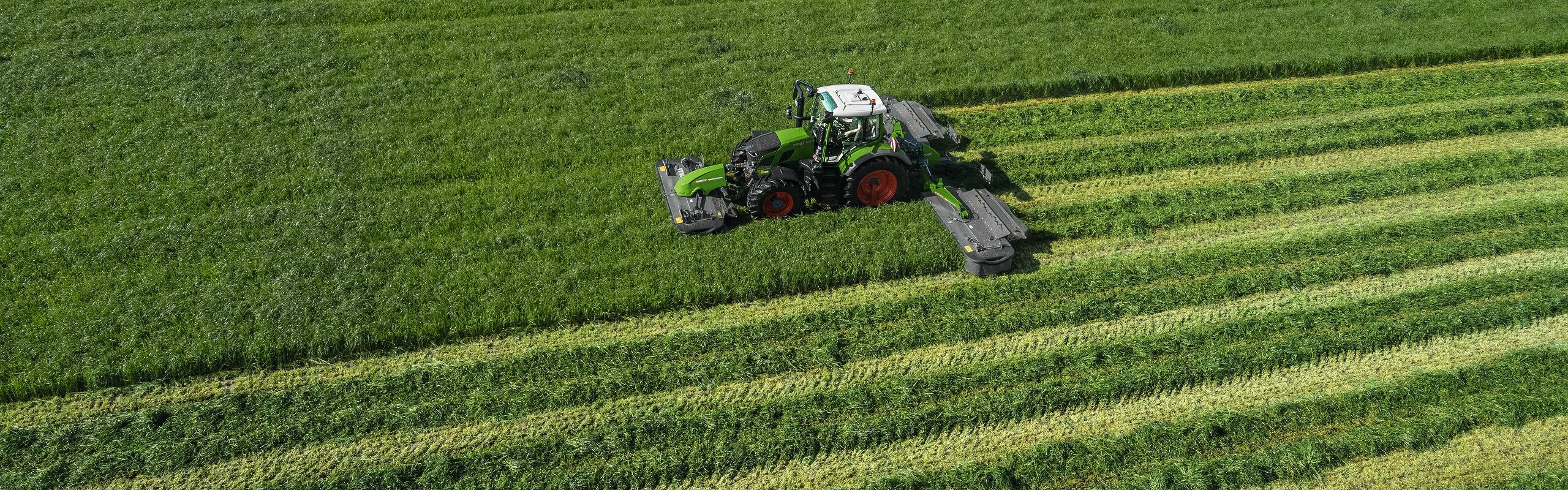 Vogelperspektive des grünen Traktors Fendt 600 Vario im Einsatz mit dem grünen Mähwerk Fendt Slicer auf einer großen, grünen Wiese.