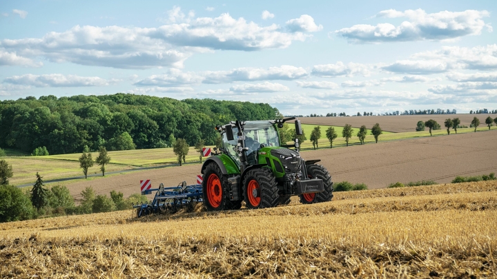 Ein grüner Fendt Traktor mit roten Felgen bei der Bodenbearbeitung auf einem abgeernteten Feld mit sanfter Hügellandschaft im Hintergrund