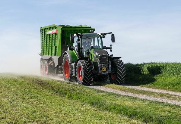 Der grüne Fendt 600 Vario Traktor mit roten Felgen und dem grünen Ladewagen Fendt Tigo fährt auf einer kleinen Feldstraße mit einer grünen Wiese im Vorder- und Hintergrund.