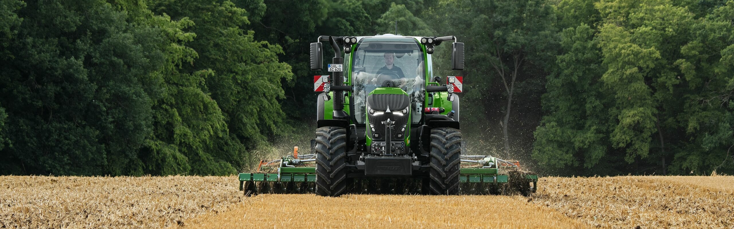 Der grüne Fendt 600 Vario ist im Einsatz mit einem Anbaugerät auf einem braunen Feld. Im Hintergrund ist ein dunkelgrüner Wald und ein wolkenbehangener Himmel.
