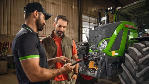 Fendt Service Ein Service Mitarbeiter mit schwarzer Cappy steht neben einem Mann und hält ein Tablet in der Hand, rechts ist ein Fendt Traktor zu sehen.