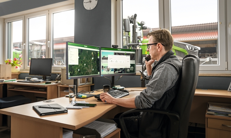 Ein Mann sitzt an einem Schreibtisch und schaut auf Computerbildschirme. Durch das Fenster kann man die Front eines grünen Fendt Traktors sehen.
