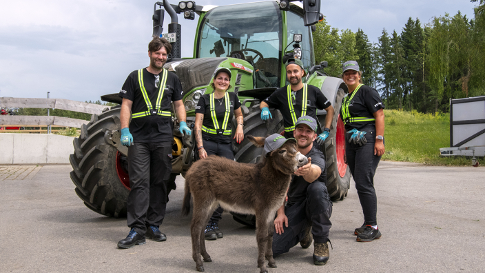 5 Personen, davon 2 Frauen stehen vor einem grünen Fendt Traktor und tragen Arbeitsschutzkleidung. Vor ihnen hockt ein Mann und füttert einen Esel mit Fendt Cap