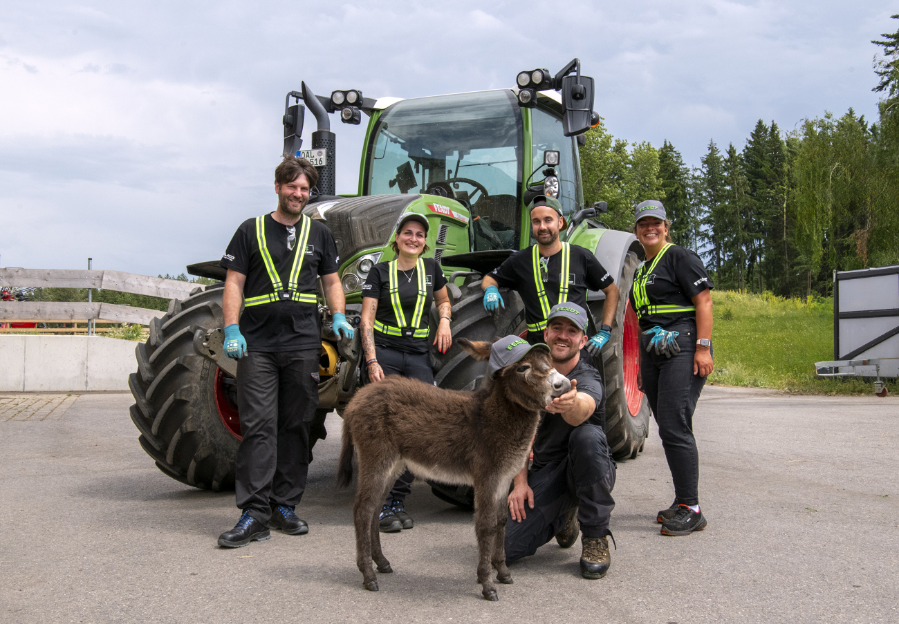 5 Personen, davon 2 Frauen stehen vor einem grünen Fendt Traktor und tragen Arbeitsschutzkleidung. Vor ihnen hockt ein Mann und füttert einen Esel mit Fendt Cap