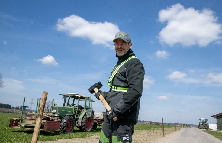 Ein Mann in Arbeitsschutzkleidung steht lächelnd vor einem Holzpfahl und hält einen Vorschlaghammer in der Hand. Links von ihm im Hintergrund steht ein grüner Fendt Traktor, auf dem weitere Pfähle liegen.