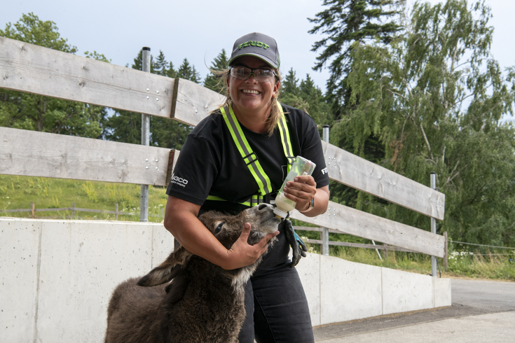 Eine Frau in Arbeitsschutzkleidung lächelt in die Kamera, während sie einen Esel mit einer Flasche füttert.