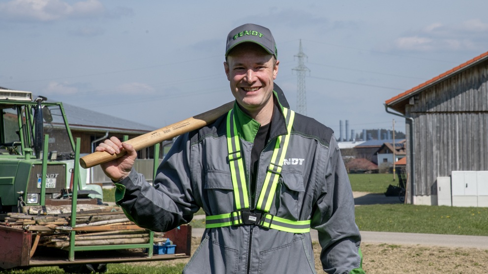 Ein Mann in Arbeitskleidung lächelt in die Kamera, während er einen Vorschlaghammer hält. Hinter ihm befindet sich ein Holzhaus (rechts) und ein grüner Fendt Traktor (links) mit Holzpfählen beladen