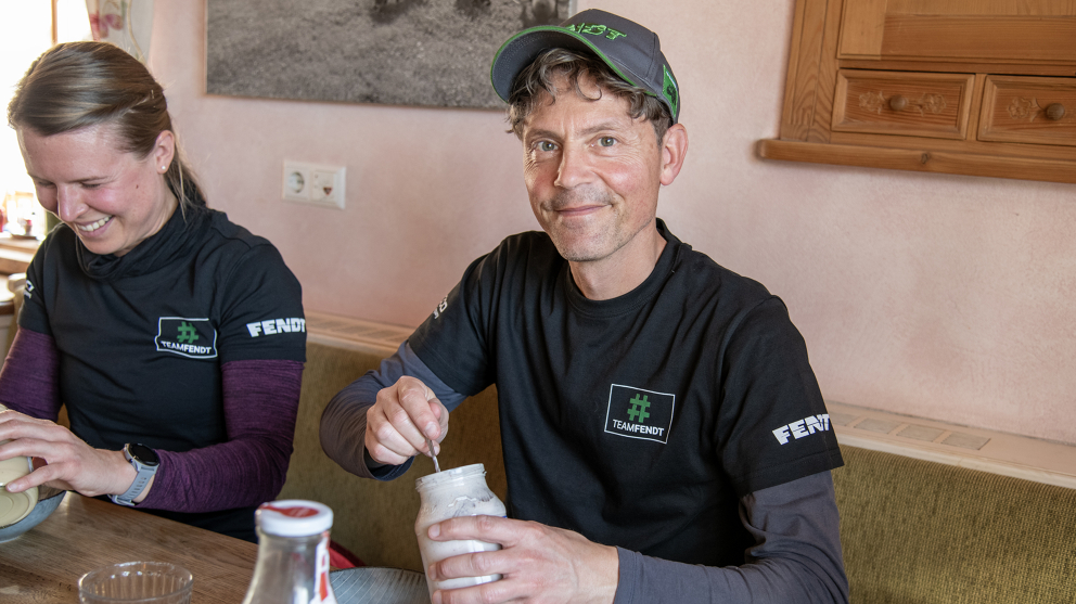 Eine Frau (links) und ein Mann (rechts) tragen Fendt Shirts und machen Mittagspause. Die Frau schaut lächelnd auf ihren Teller, während der Mann, der ein Glas Joghurt in der Hand hält, lächeln in die Kamera schaut.