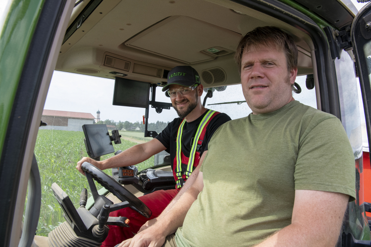 2 Männer sitzen in einem Traktor, dessen Innenraum zu sehen ist, und schauen in die Kamera. Einer sitzt am Lenkrad.