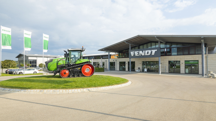 Rechts ein großes Gebäude mit einem Fendt Schild, links daneben steht ein Fendt Traktor mit weiß/grüne Fendt Fahnen.