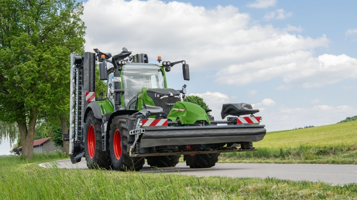 Der grüne Fendt 700 Vario mit Front- und Heckmähwerken fährt auf einer Landstraße umgeben von Wiesen. Im Hintergrund ist ein Baum und der blaue, leicht bewölkte Himmel zu sehen.