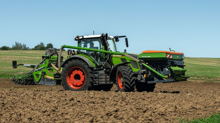 Der grüne Fendt 600 Vario mit roten Felgen und einer Drillmaschine als Anbaugerät fährt auf einem Acker. Im Hintergrund ist eine grüne Wiese und ein blauer Himmel zu sehen.