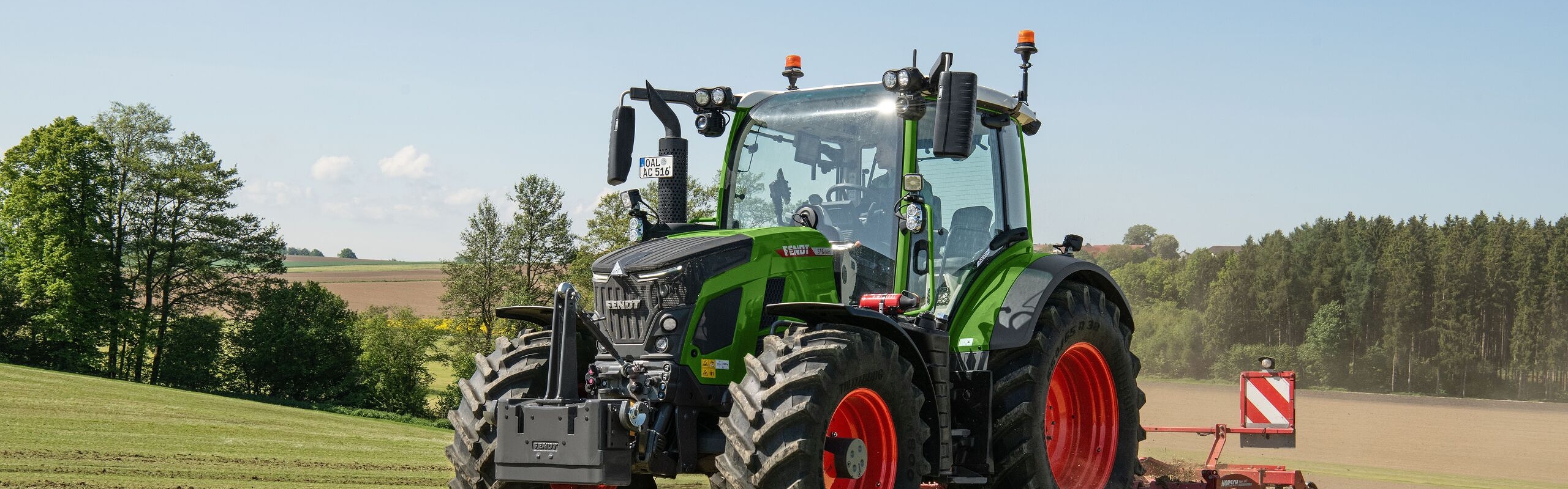 Ein Fendt 500 Vario Gen4 Traktor in grün mit roten Felgen fährt auf einer hellgrünen Wiese. Hinten ist ein rotes Bodenbearbeitungsgerät mit Warntafel angehängt. Im Hintergrund sind Felder, ein Waldrand und blauer Himmel mit wenigen Wolken zu sehen.
