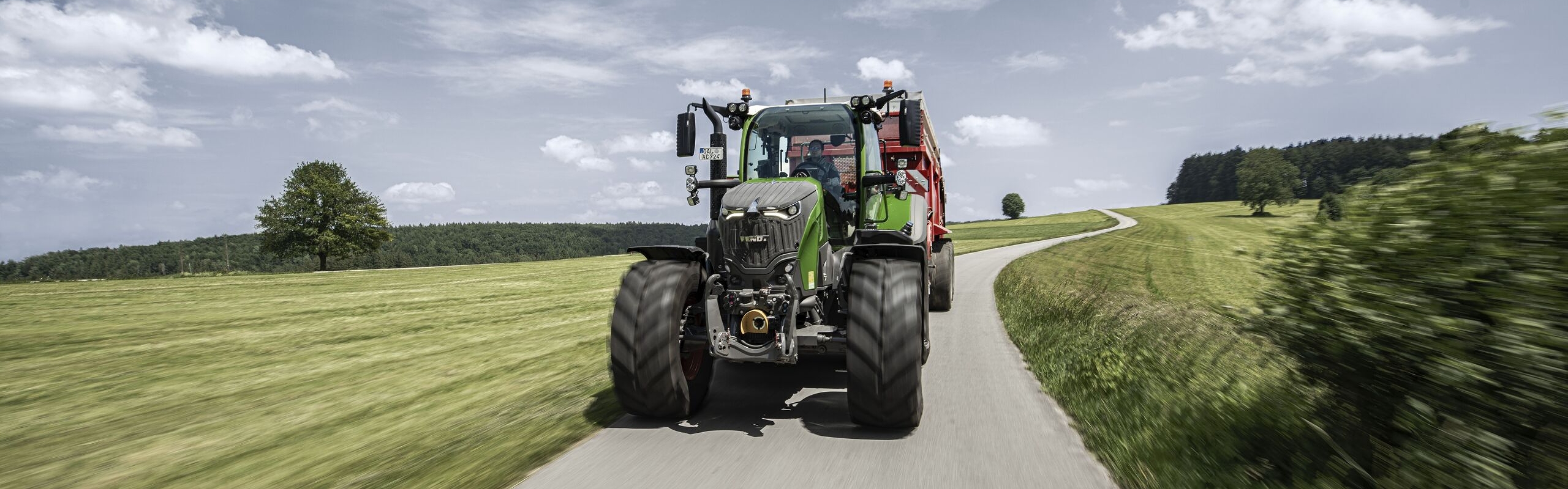 Ein Fendt 700 Vario Gen7.1 fährt mit angebautem Gerät auf einer schmalen Landstraße durch eine grüne Hügellandschaft, bei leicht bewölktem Himmel.